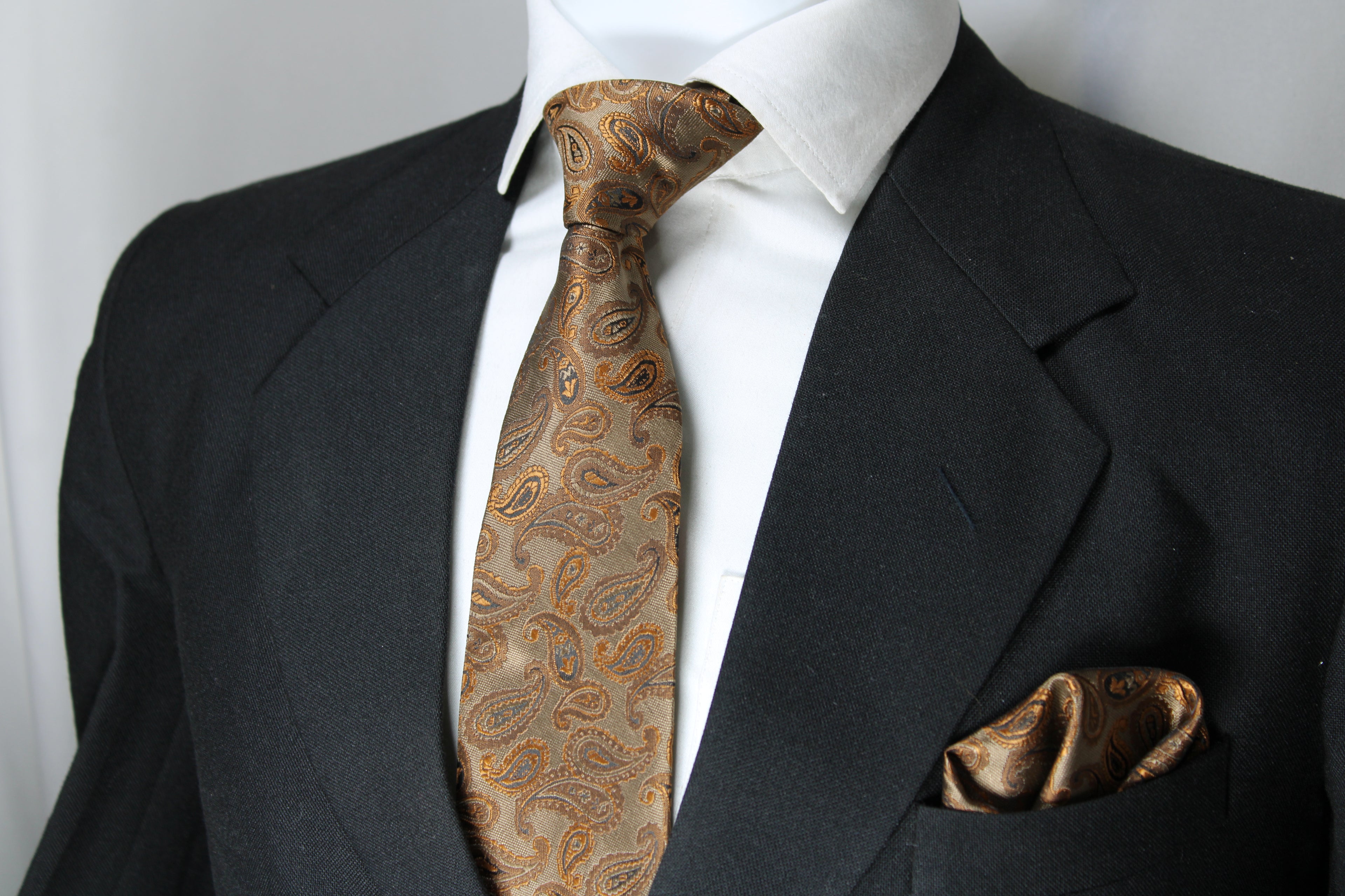 A light brown and gold paisley necktie paired with a matching pocket square displayed on a mannequin wearing a black suit and a white shirt.
