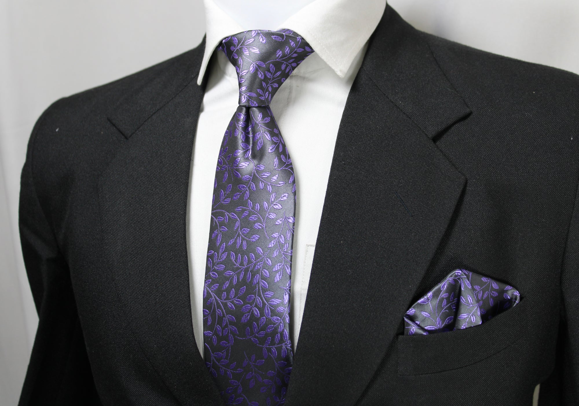A mannequin wearing a black suit, displaying a gray and purple floral tie and a matching pocket square.