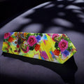 A yellow necktie with a floral pattern laid out flat on a dark surface, with the shadow of a plant leaf across the image.