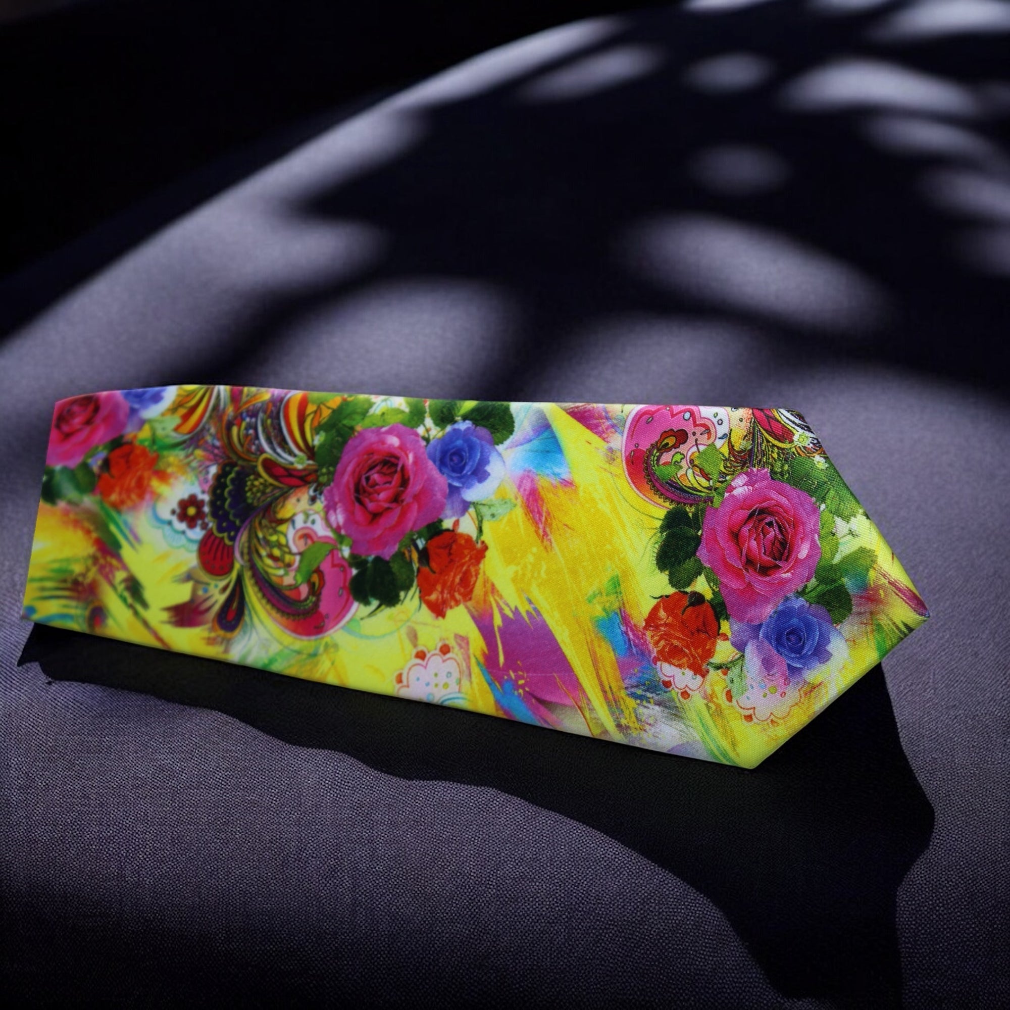 A yellow necktie with a floral pattern laid out flat on a dark surface, with the shadow of a plant leaf across the image.