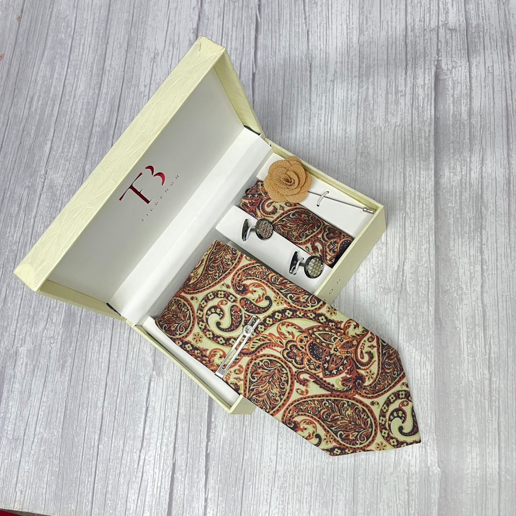 A beige paisley printed silk tie combo set displayed in an open white box, including a necktie, pocket square, lapel flower pin, cufflinks, and a brass tie pin.