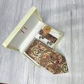 A beige paisley printed silk tie combo set displayed in an open white box, including a necktie, pocket square, lapel flower pin, cufflinks, and a brass tie pin.