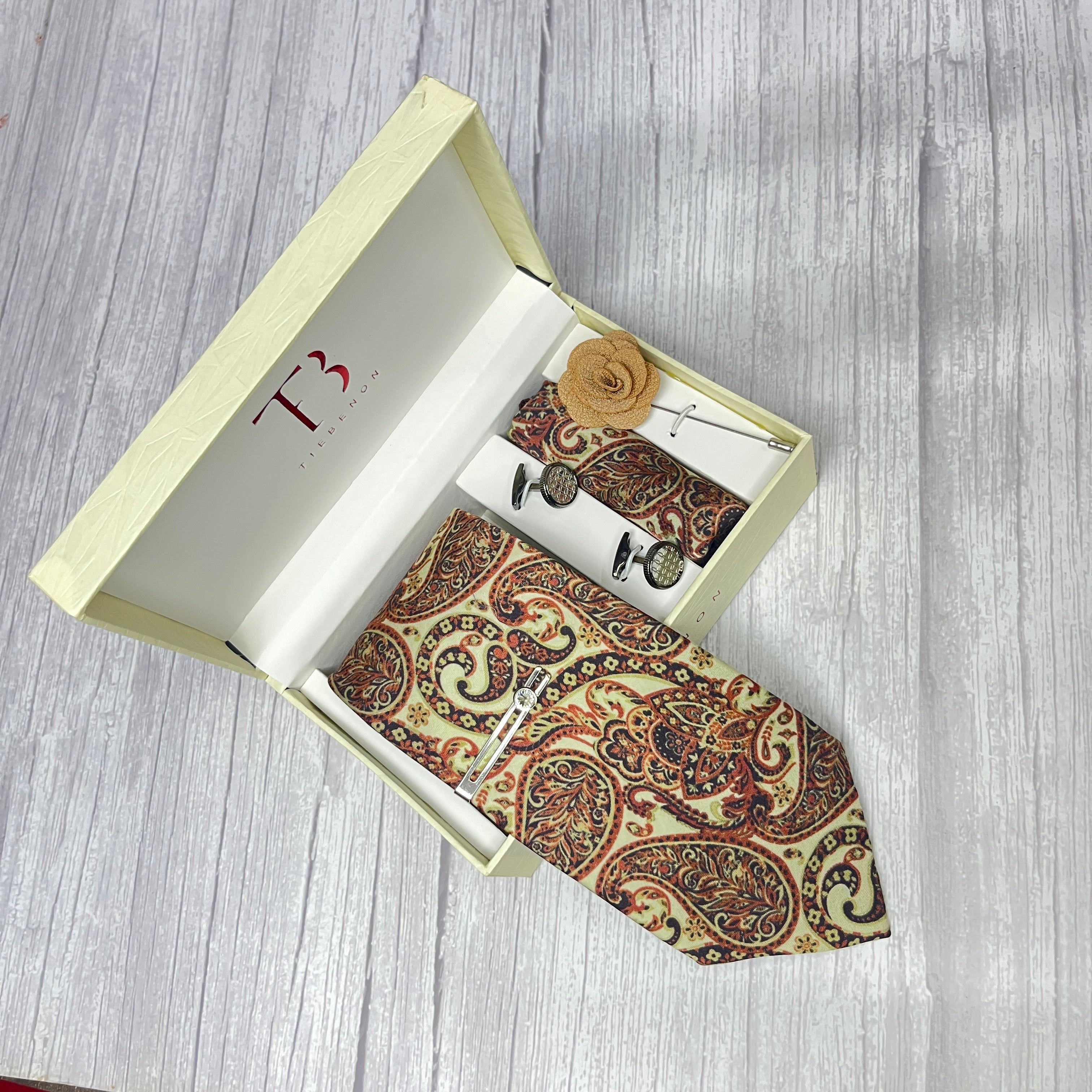 A beige paisley printed silk tie combo set displayed in an open white box, including a necktie, pocket square, lapel flower pin, cufflinks, and a brass tie pin.