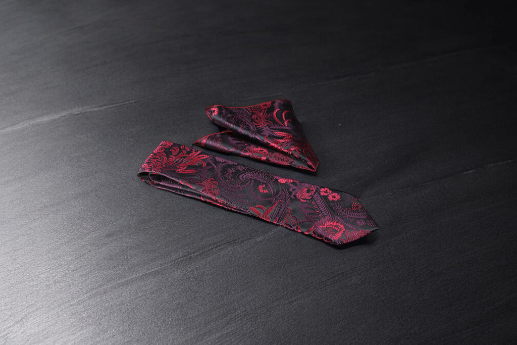 A black and maroon floral tie and pocket square set, laid out on a dark surface.