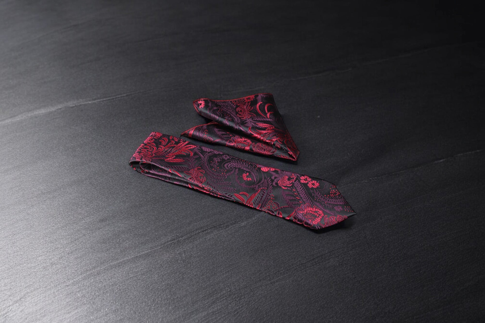 A black and maroon floral tie and pocket square set, laid out on a dark surface.