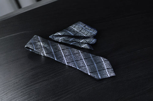 A silver and black floral plaid tie and pocket square set, laid out on a dark surface.