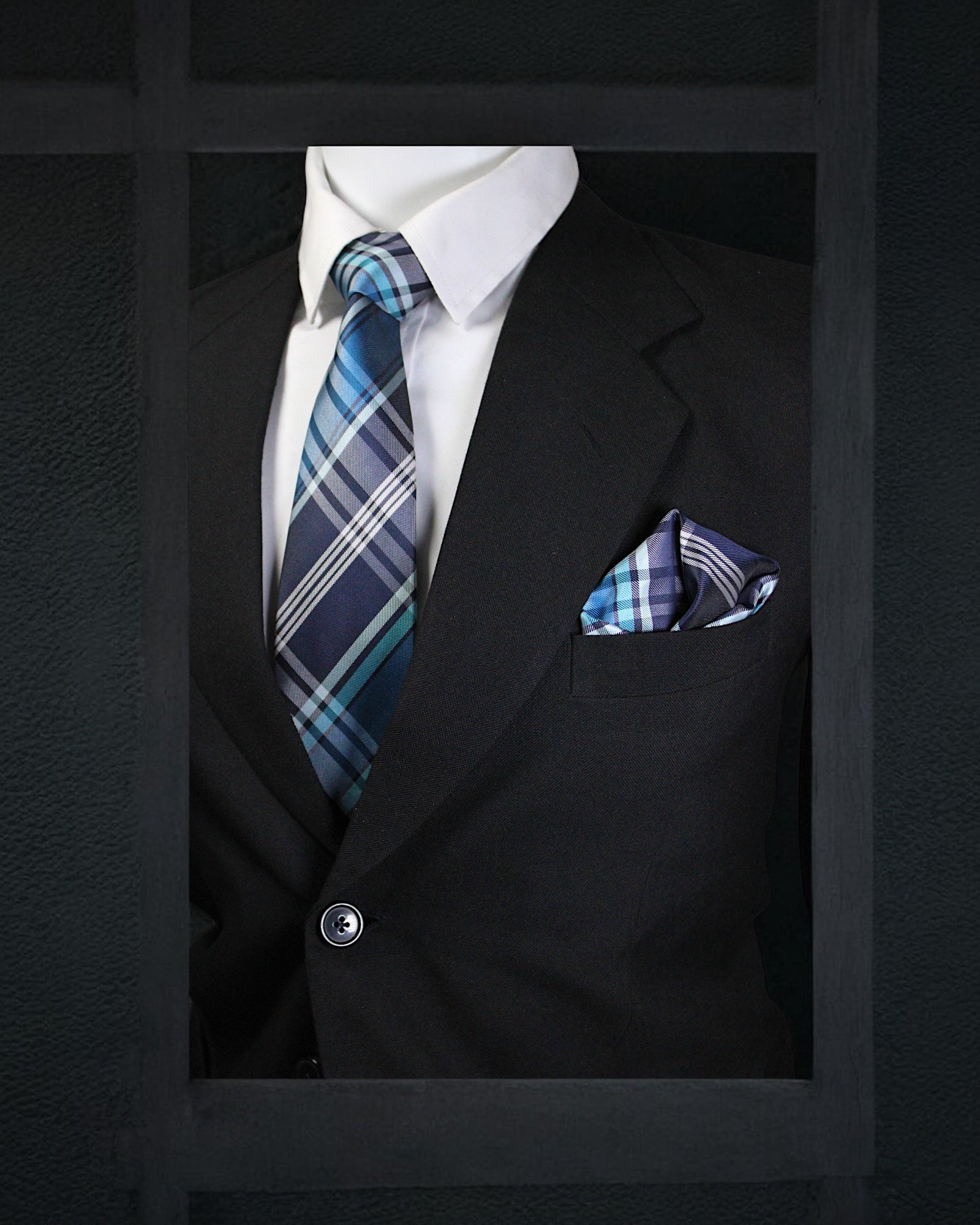Black suit with white shirt, blue plaid tie, and matching pocket square on a dark background