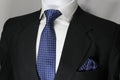 A blue multicolor geometric tie with a matching pocket square displayed on a mannequin wearing a black suit and a white shirt.