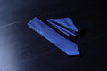 A blue and navy geometric patterned tie and pocket square set laid out on a dark surface.