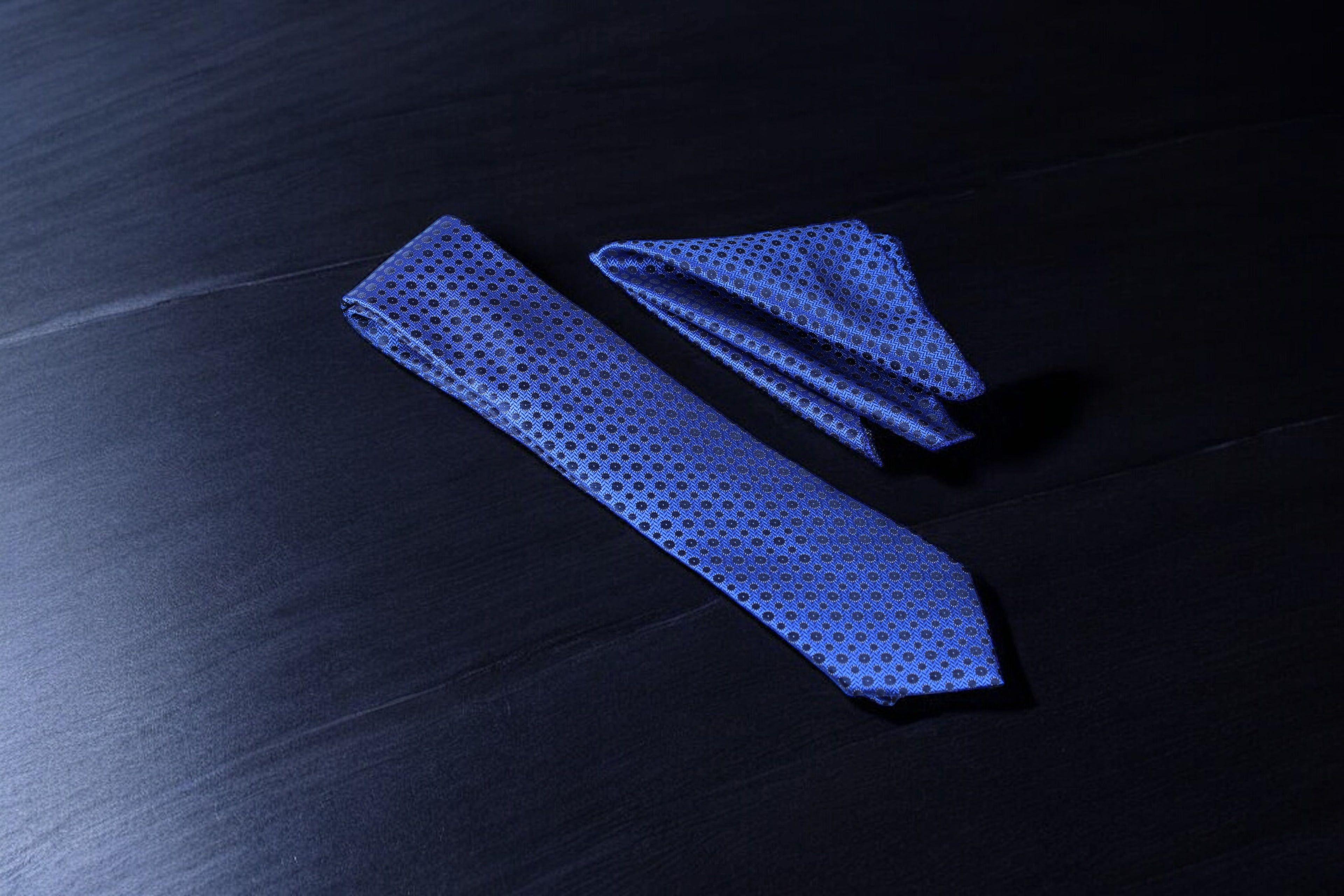 A blue and navy geometric patterned tie and pocket square set laid out on a dark surface.