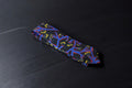 A black necktie with a bold geometric botanical pattern, laid out on a dark surface.