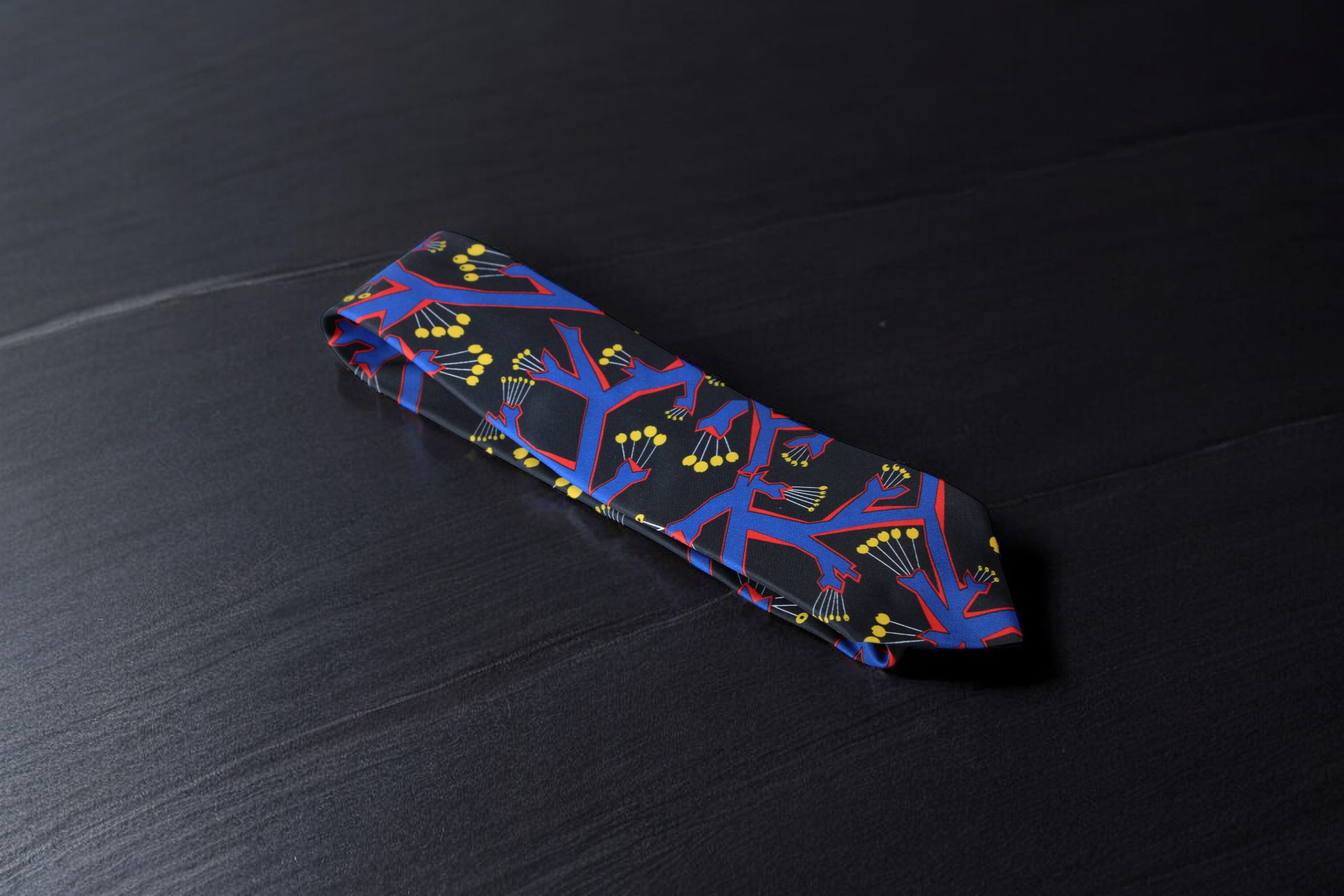 A black necktie with a bold geometric botanical pattern, laid out on a dark surface.