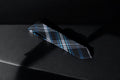 A checkered tie with navy, blue, and silver colors, lying flat on a dark surface.