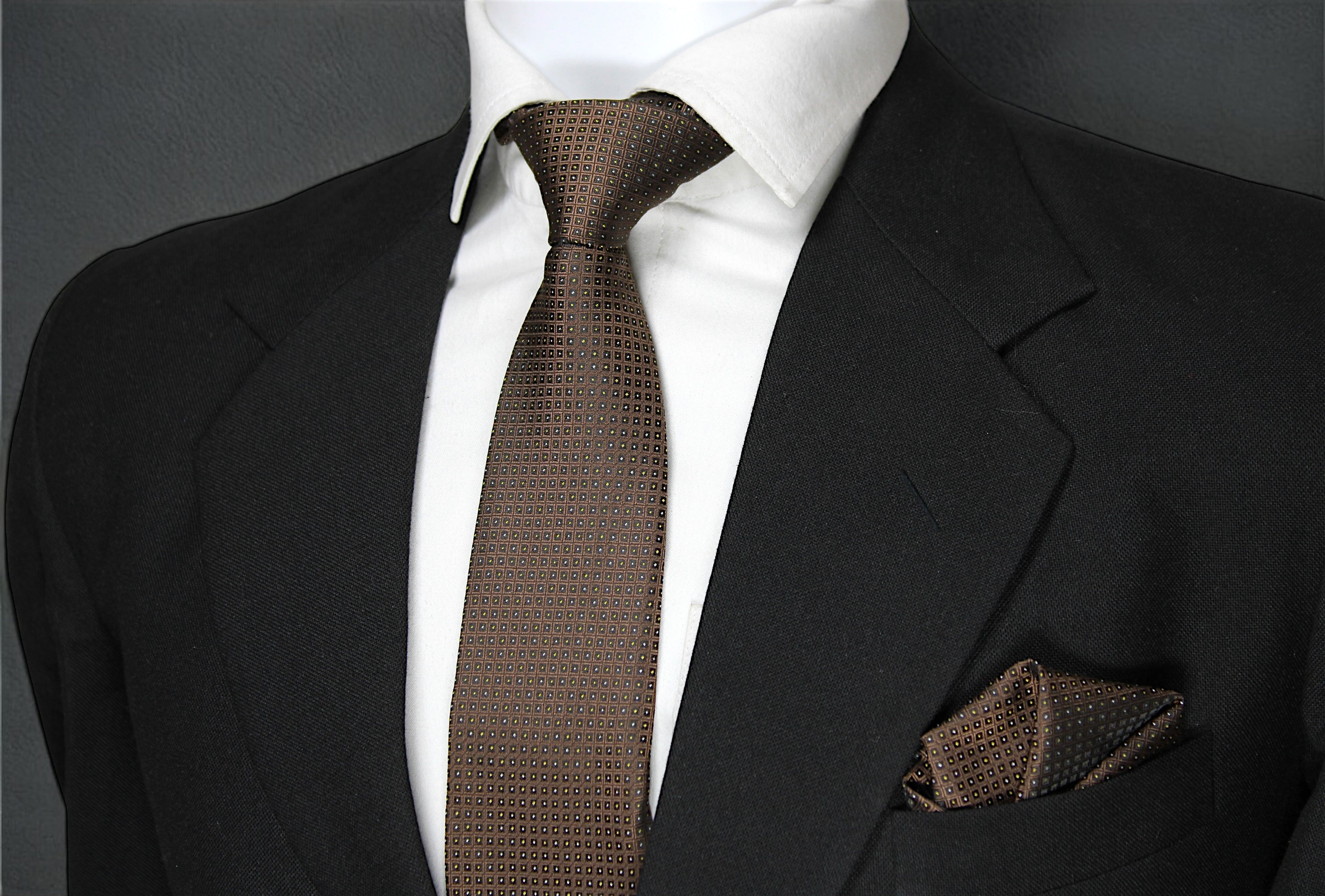 A brown geometric patterned tie and pocket square displayed on a mannequin wearing a black suit and a white shirt.