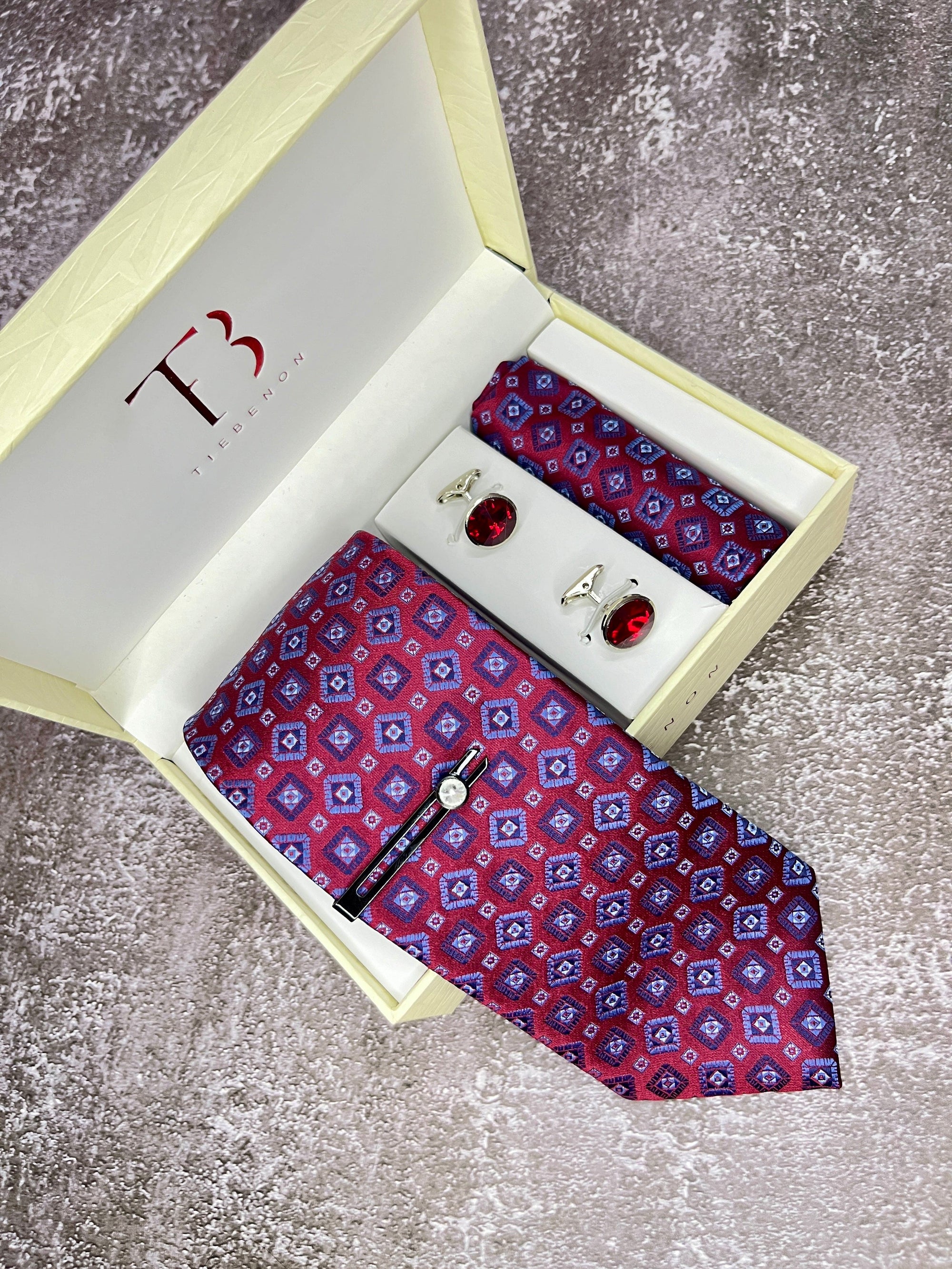A burgundy geometric patterned tie with a matching pocket square and cufflinks, presented in an open white box with a branded letter 'T' on the top.
