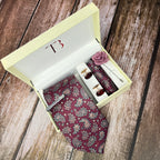 A burgundy paisley print silk tie with a matching pocket square and accessories, presented in an open white box with a yellow interior and a brand logo on the top.