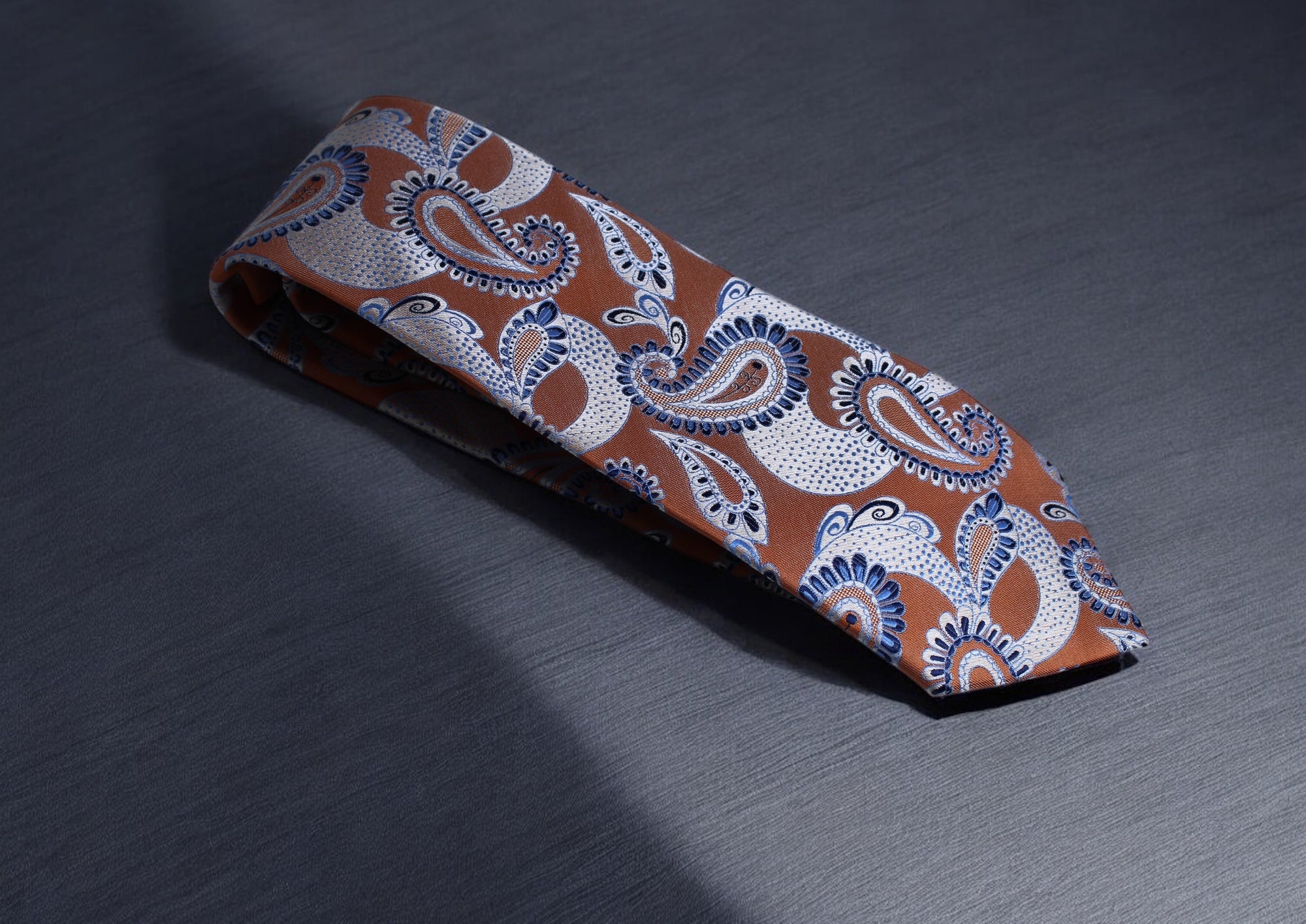 A burnt orange necktie with paisley patterns and blue and silver accents lying on a textured dark surface.