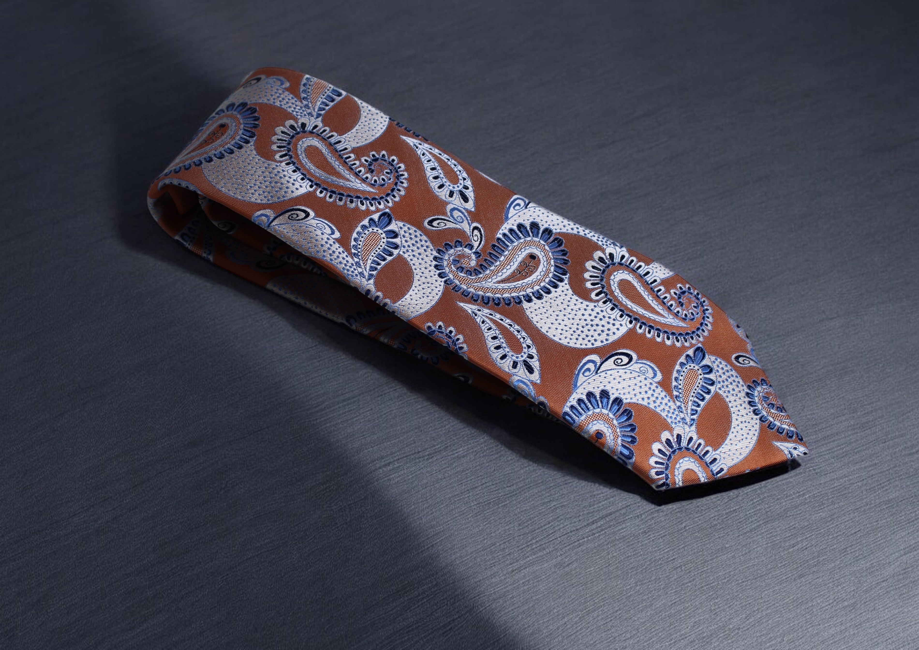 A burnt orange necktie with paisley patterns and blue and silver accents lying on a textured dark surface.