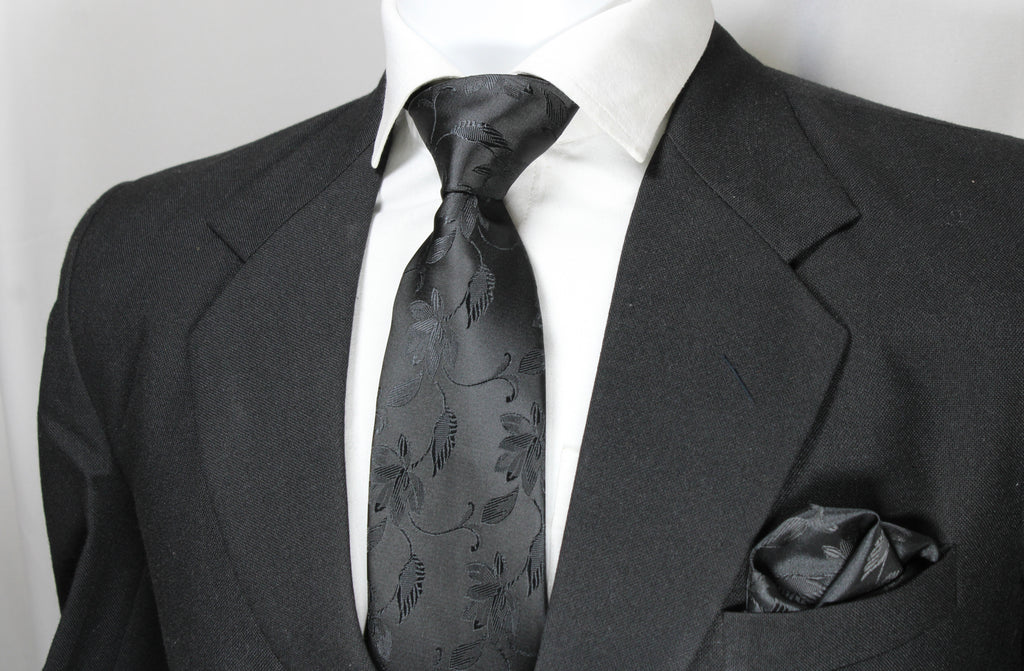 A black floral tie and pocket square set displayed on a mannequin wearing a black suit and white shirt.