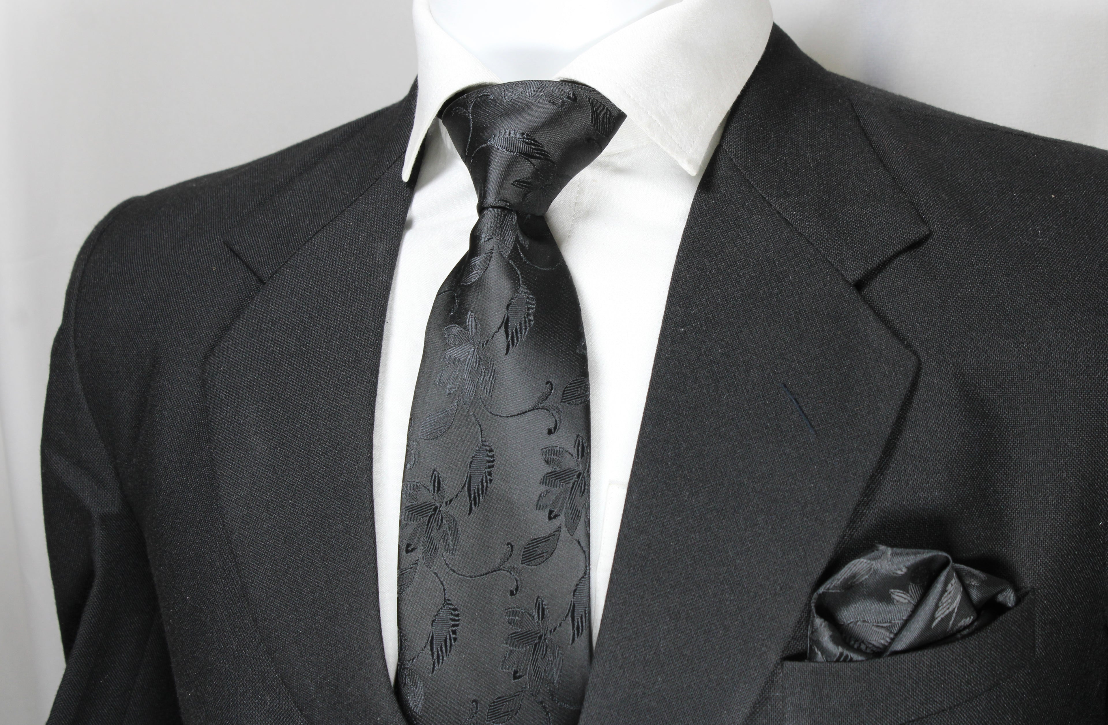 A black floral tie and pocket square set displayed on a mannequin wearing a black suit and white shirt.