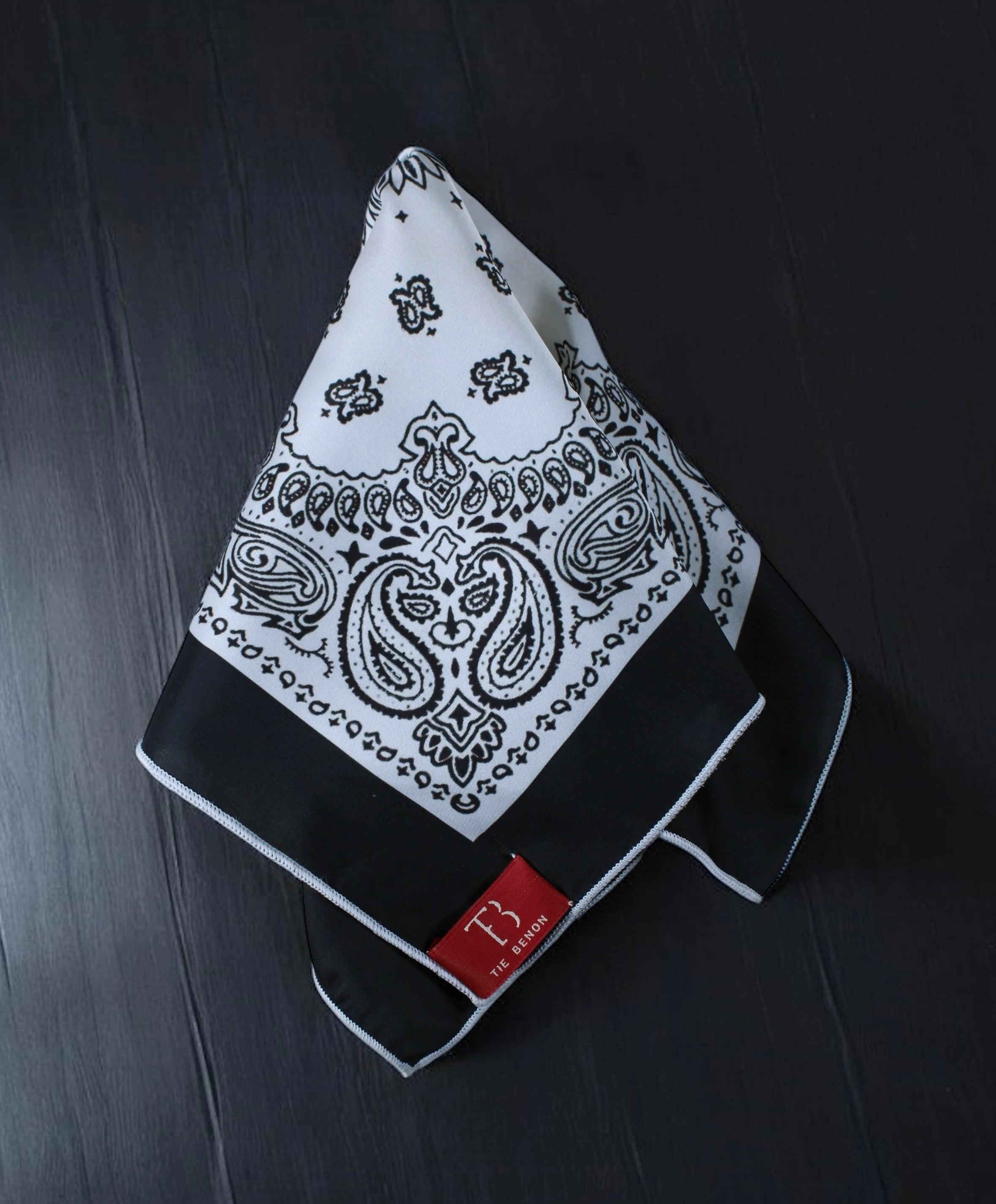 A folded black and white paisley pocket square on a dark background, with a visible brand tag.