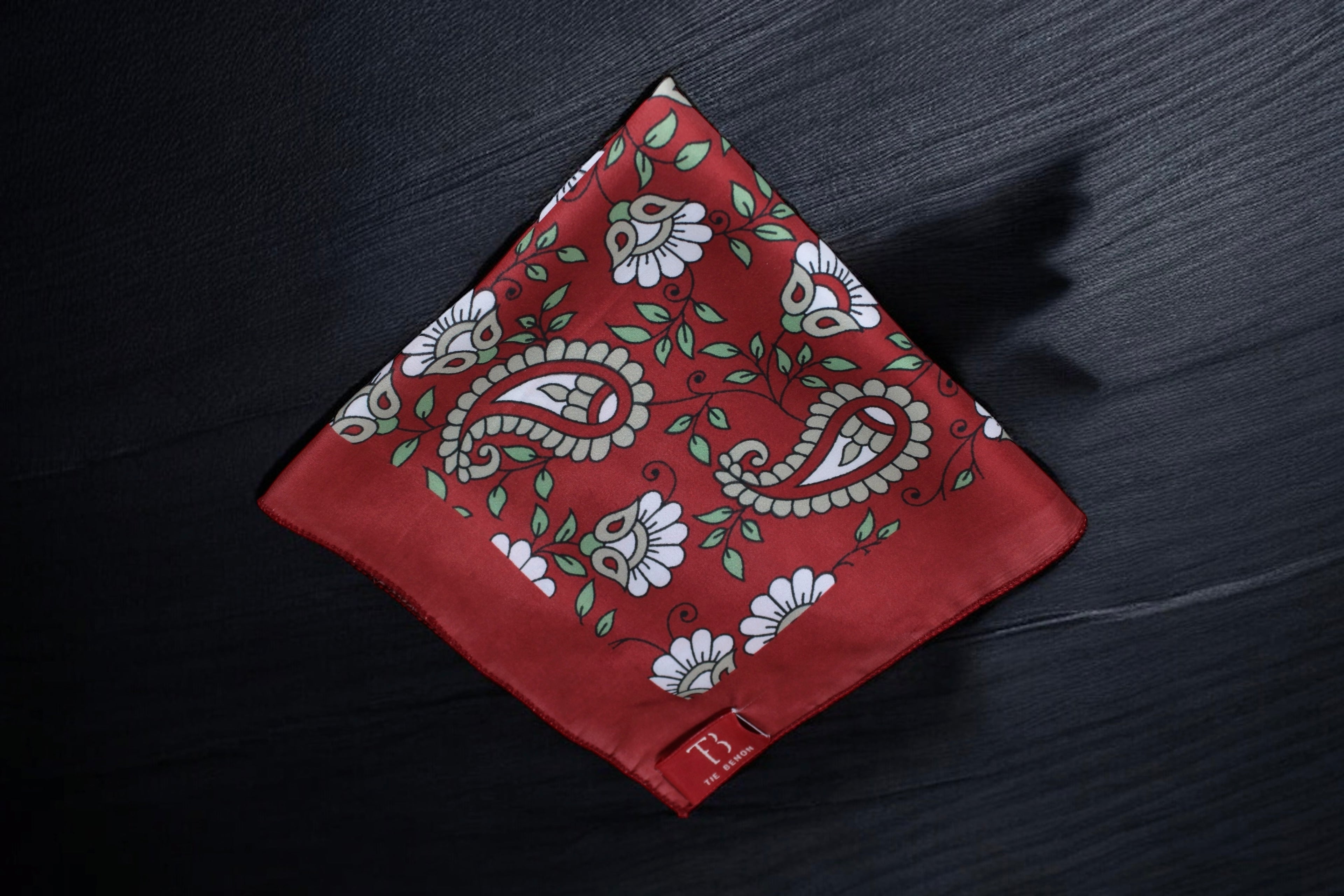 A maroon pocket square with a floral paisley pattern laid out on a dark surface.