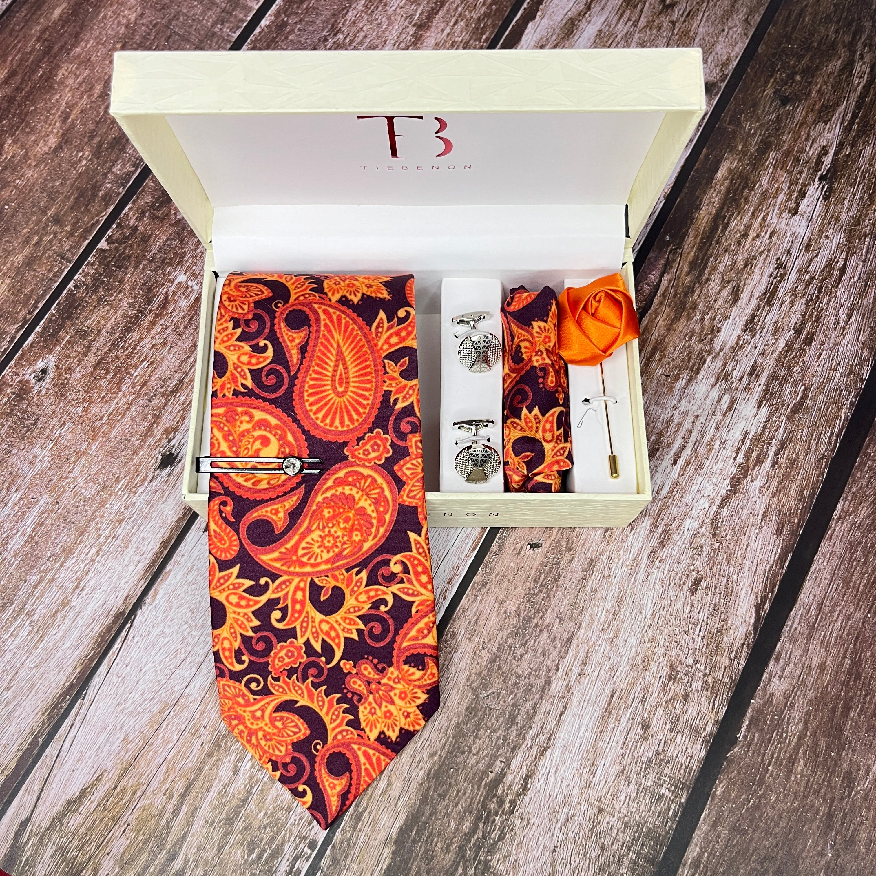 A Copper Paisley Printed Silk Tie in a box with a branded label, along with a matching pocket square, cufflinks, a lapel flower pin, and a brass tie pin.
