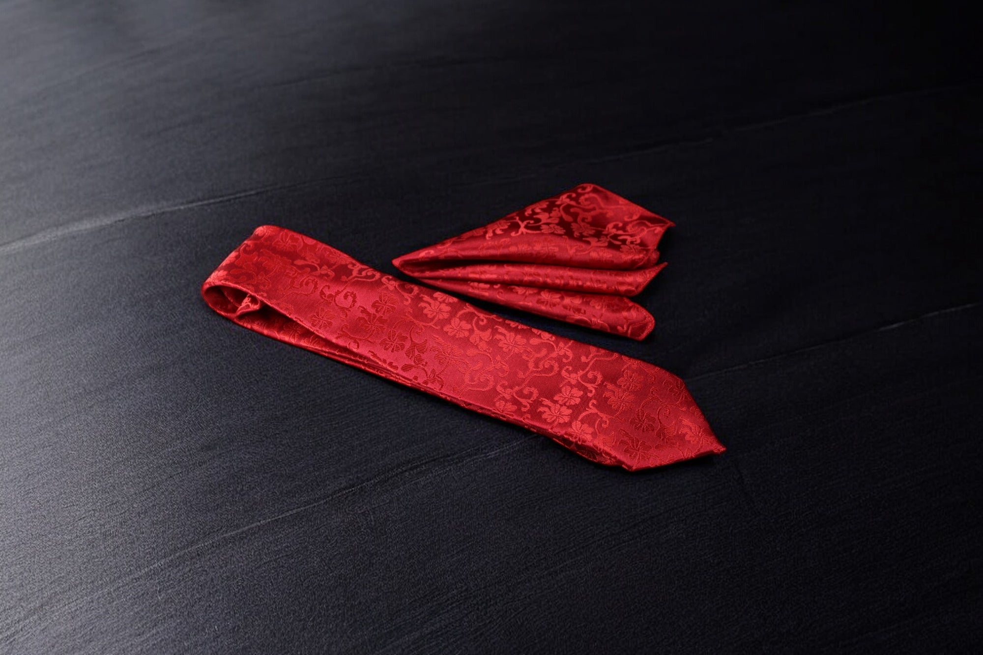 A crimson red floral tie and pocket square set laid out on a dark surface.