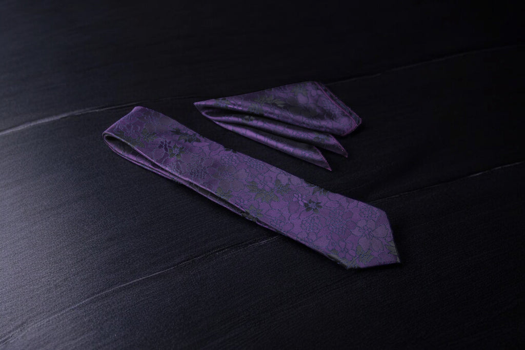 A dark purple floral tie and pocket square set, laid out on a dark surface.