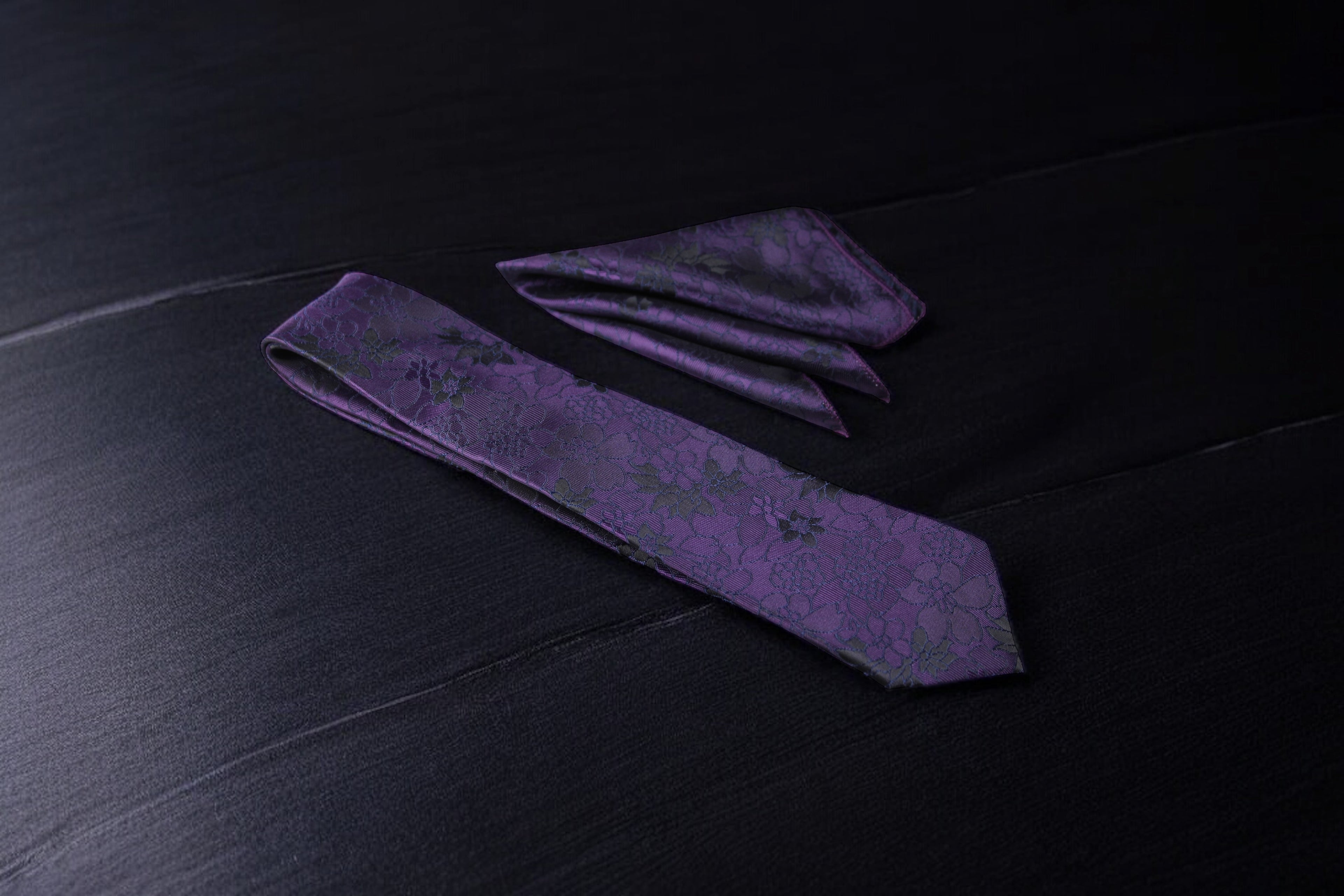 A dark purple floral tie and pocket square set, laid out on a dark surface.