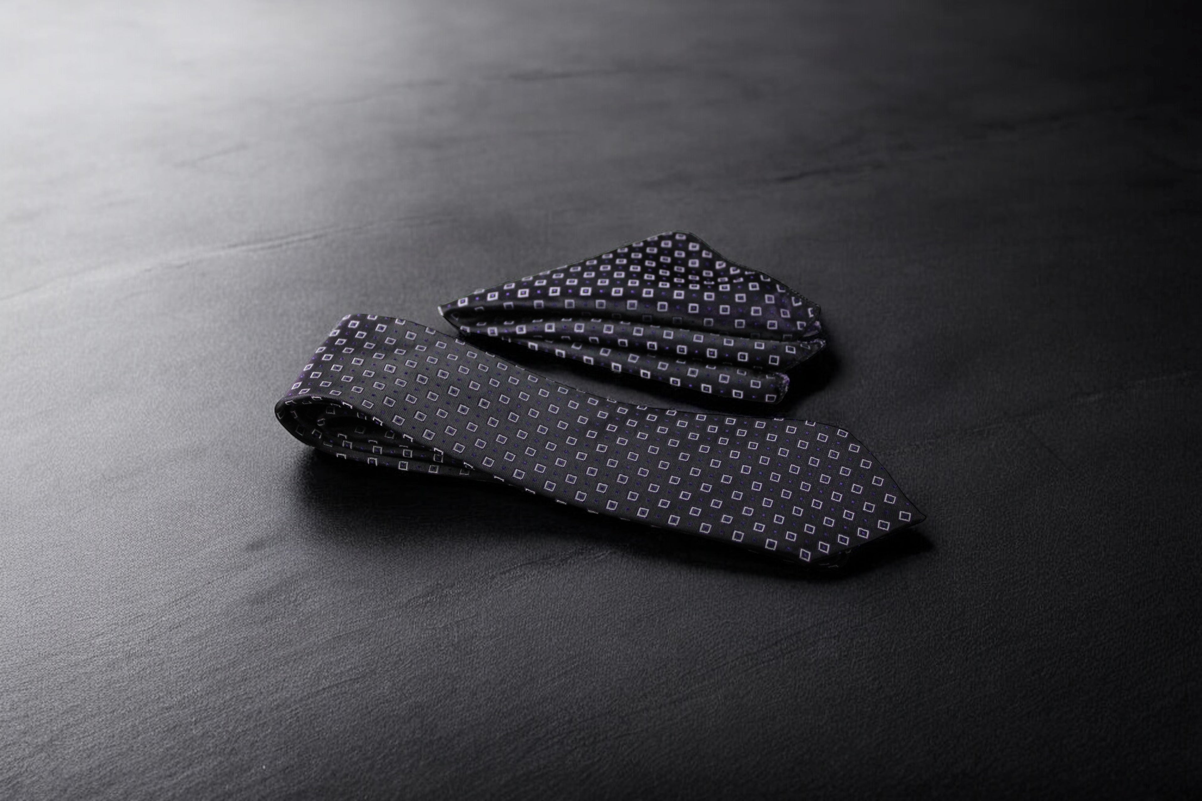 A dark purple geometric patterned tie and pocket square set laid out on a dark surface.