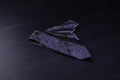 A black and purple paisley necktie and pocket square set, laid out on a dark surface.