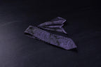 A black and purple paisley necktie and pocket square set, laid out on a dark surface.