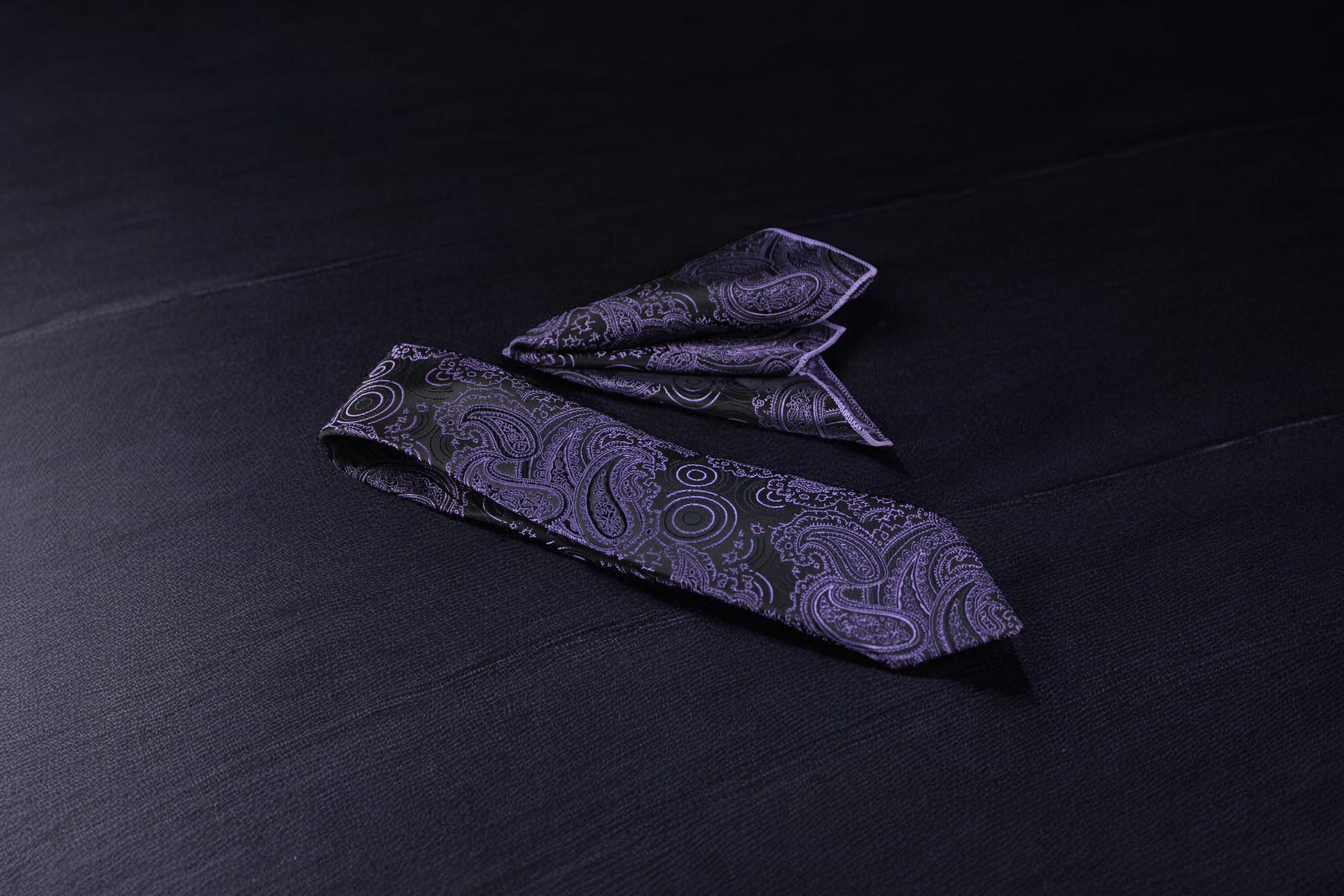 A black and purple paisley necktie and pocket square set, laid out on a dark surface.