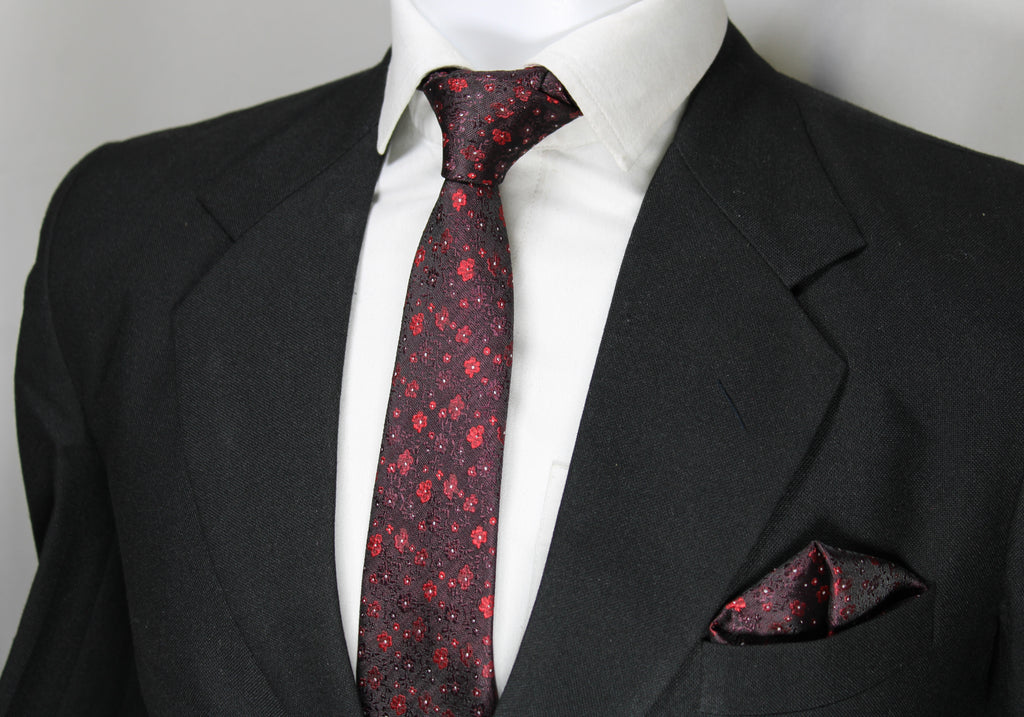 A set comprising a burgundy and red tie with a geometric pattern, displayed on a mannequin wearing a black suit and a white shirt. A matching pocket square is also visible.