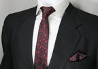 A set comprising a burgundy and red tie with a geometric pattern, displayed on a mannequin wearing a black suit and a white shirt. A matching pocket square is also visible.