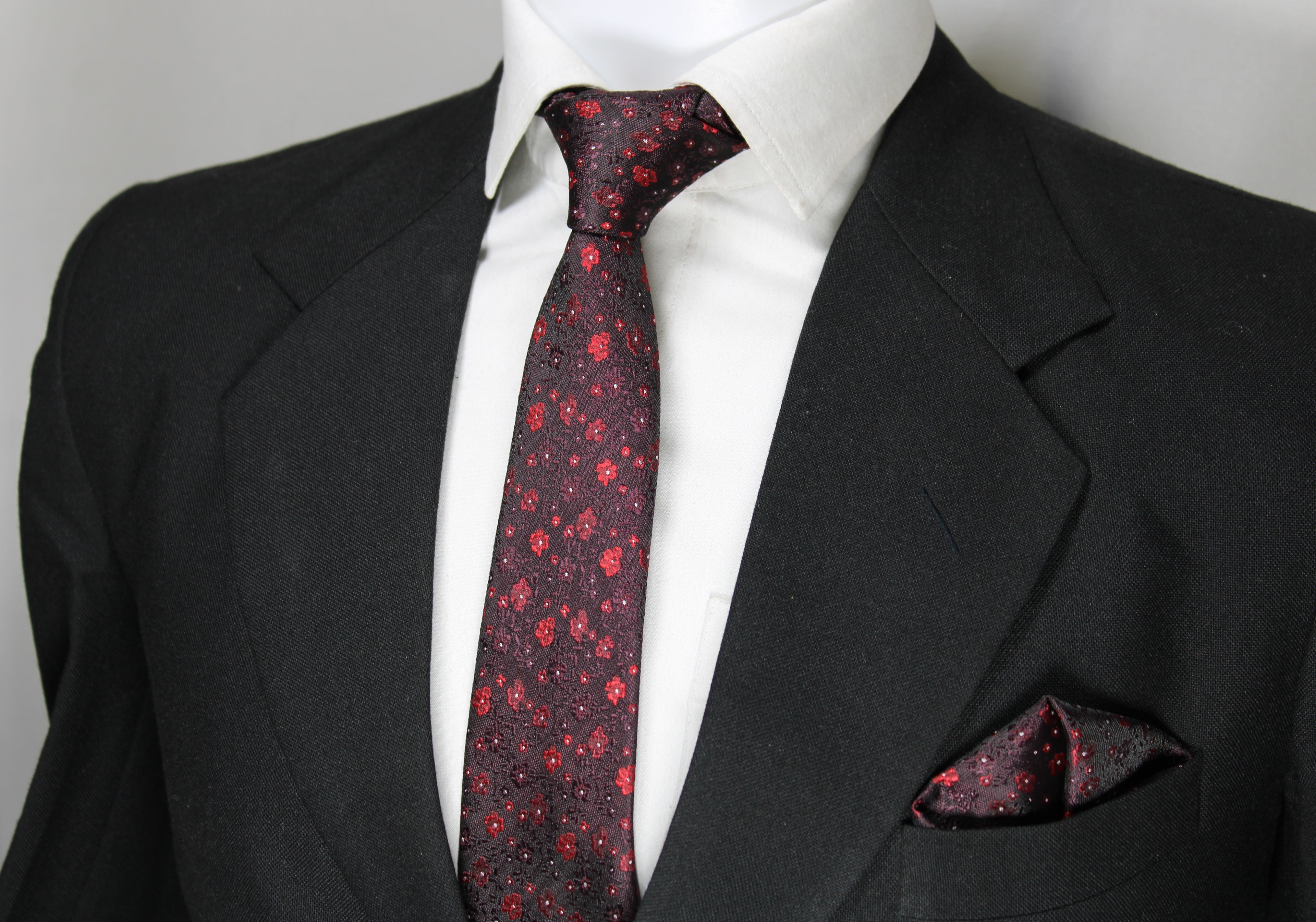 A set comprising a burgundy and red tie with a geometric pattern, displayed on a mannequin wearing a black suit and a white shirt. A matching pocket square is also visible.