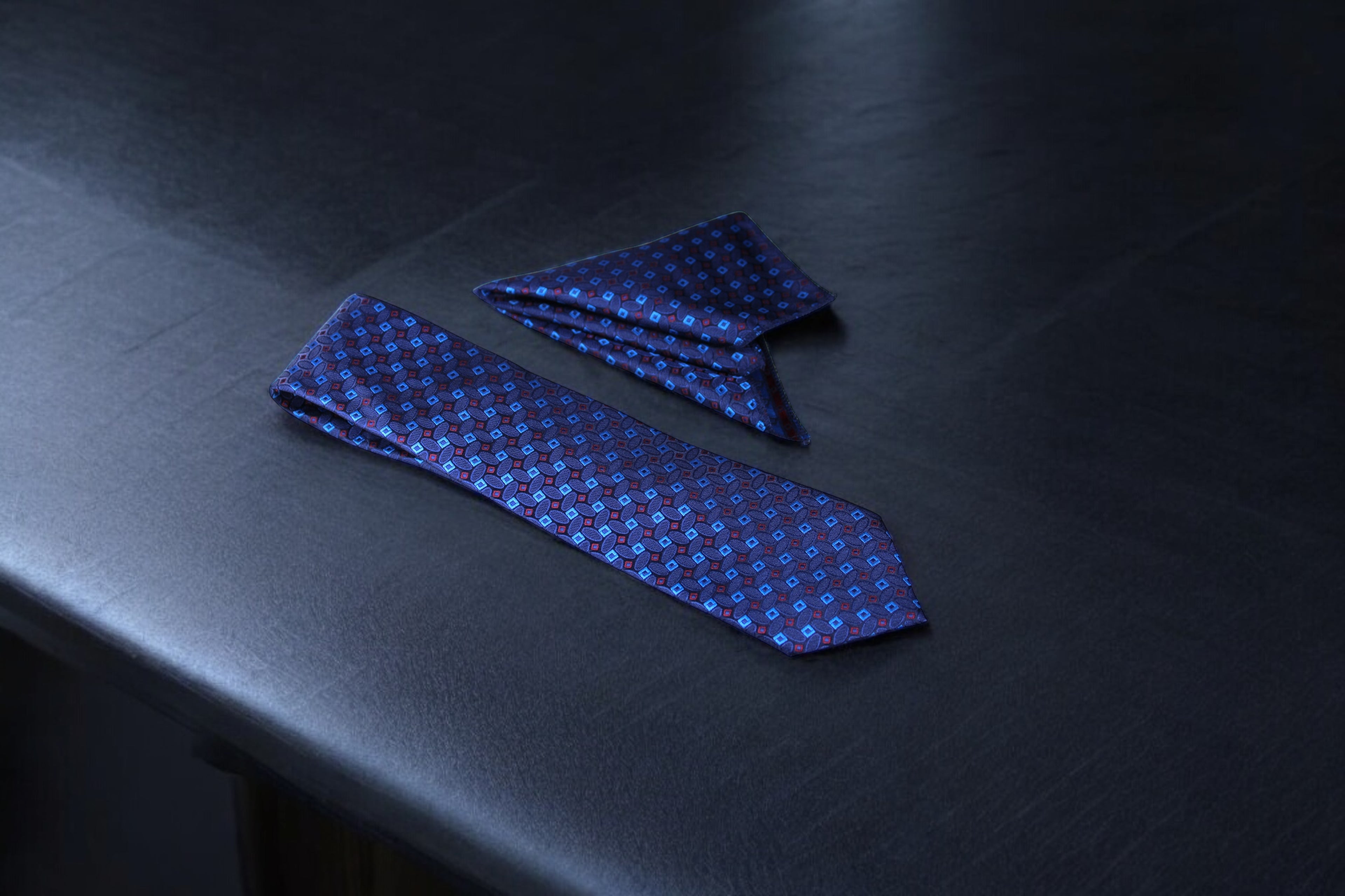 A dark blue geometric tie and a matching pocket square laid out on a dark surface.