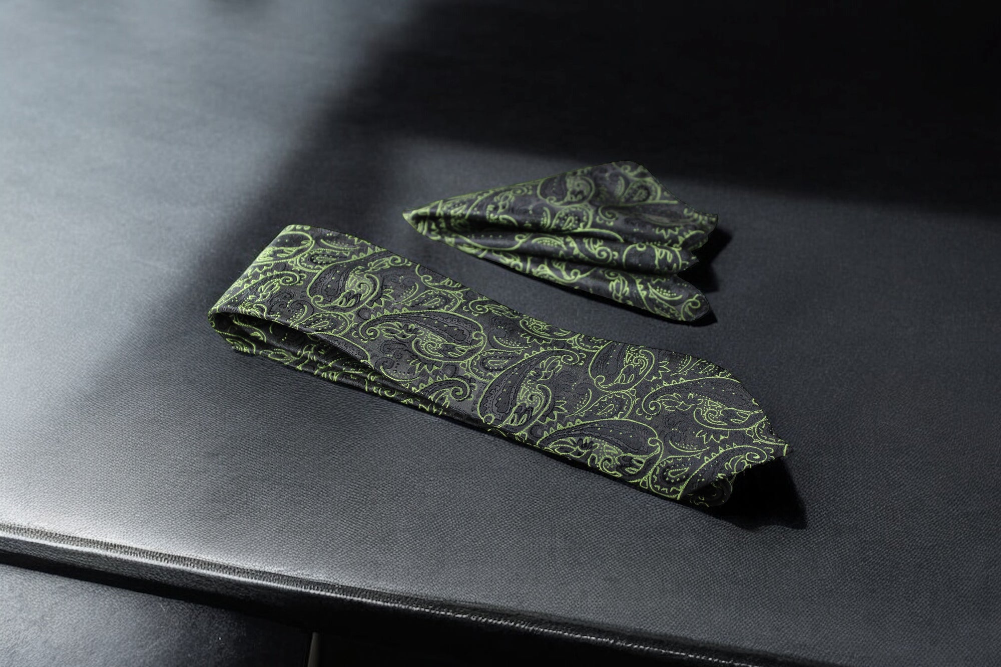 A green and black paisley tie and pocket square set laid out on a dark surface.