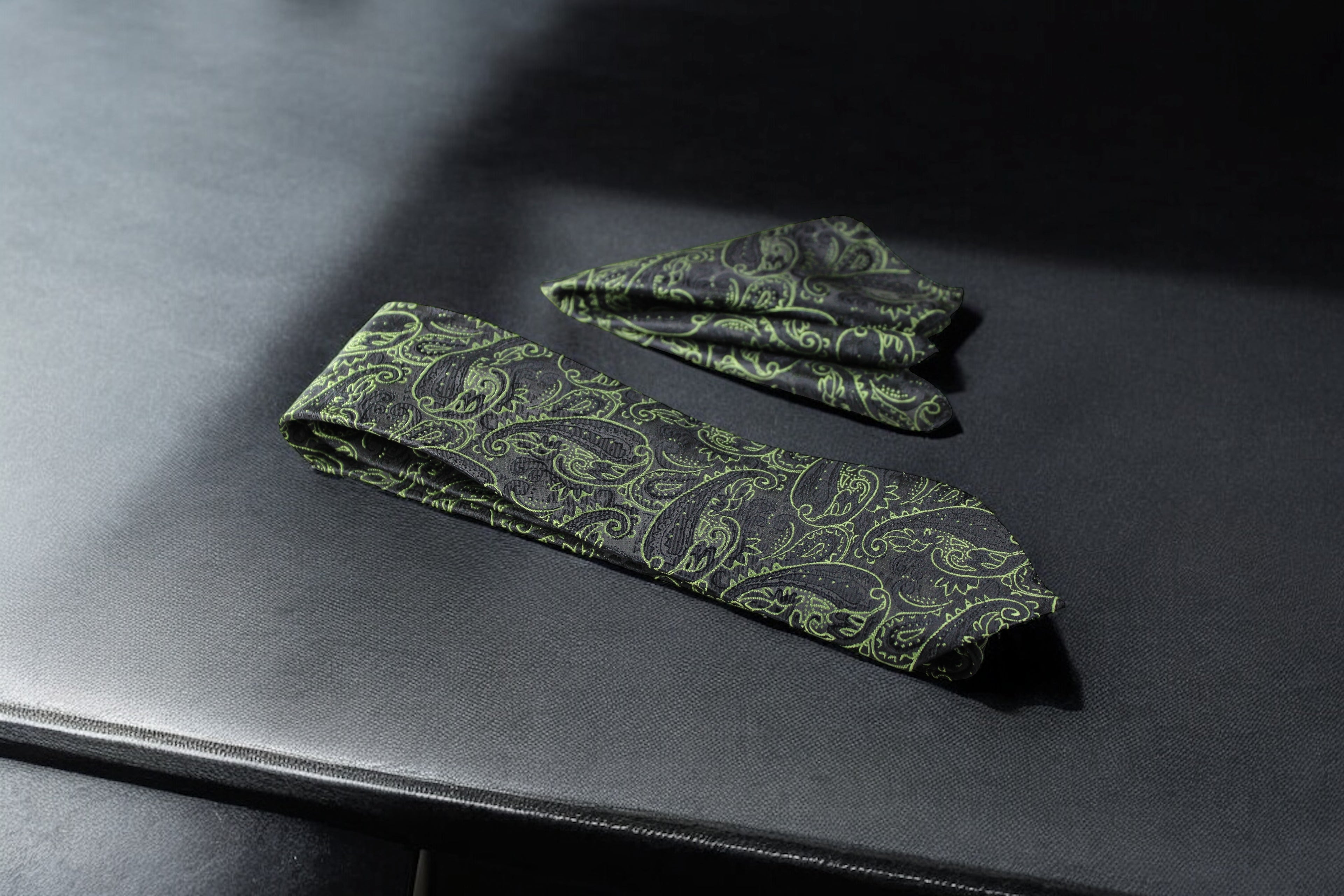 A green and black paisley tie and pocket square set laid out on a dark surface.