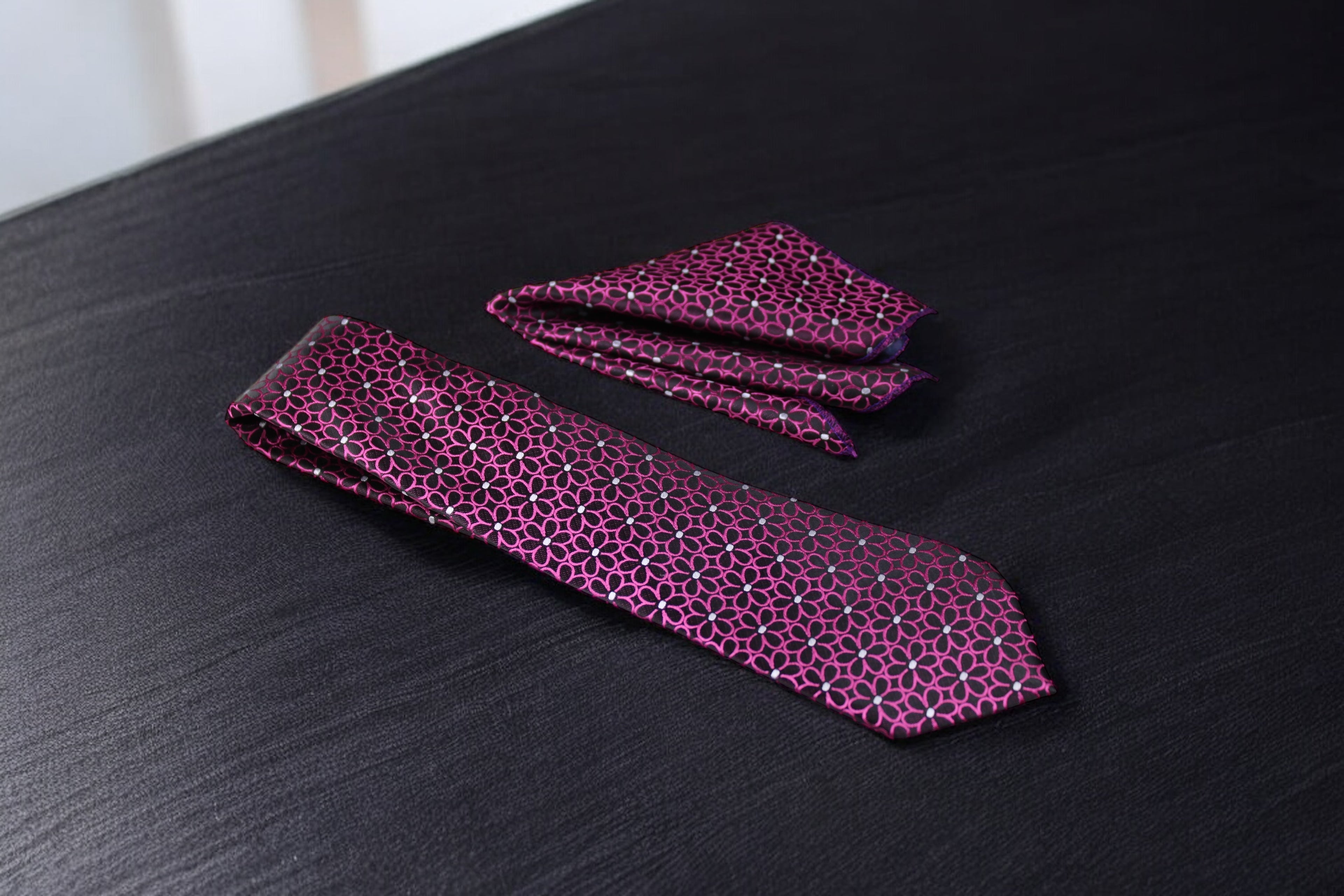 A magenta floral tie and pocket square set laid out on a dark surface.