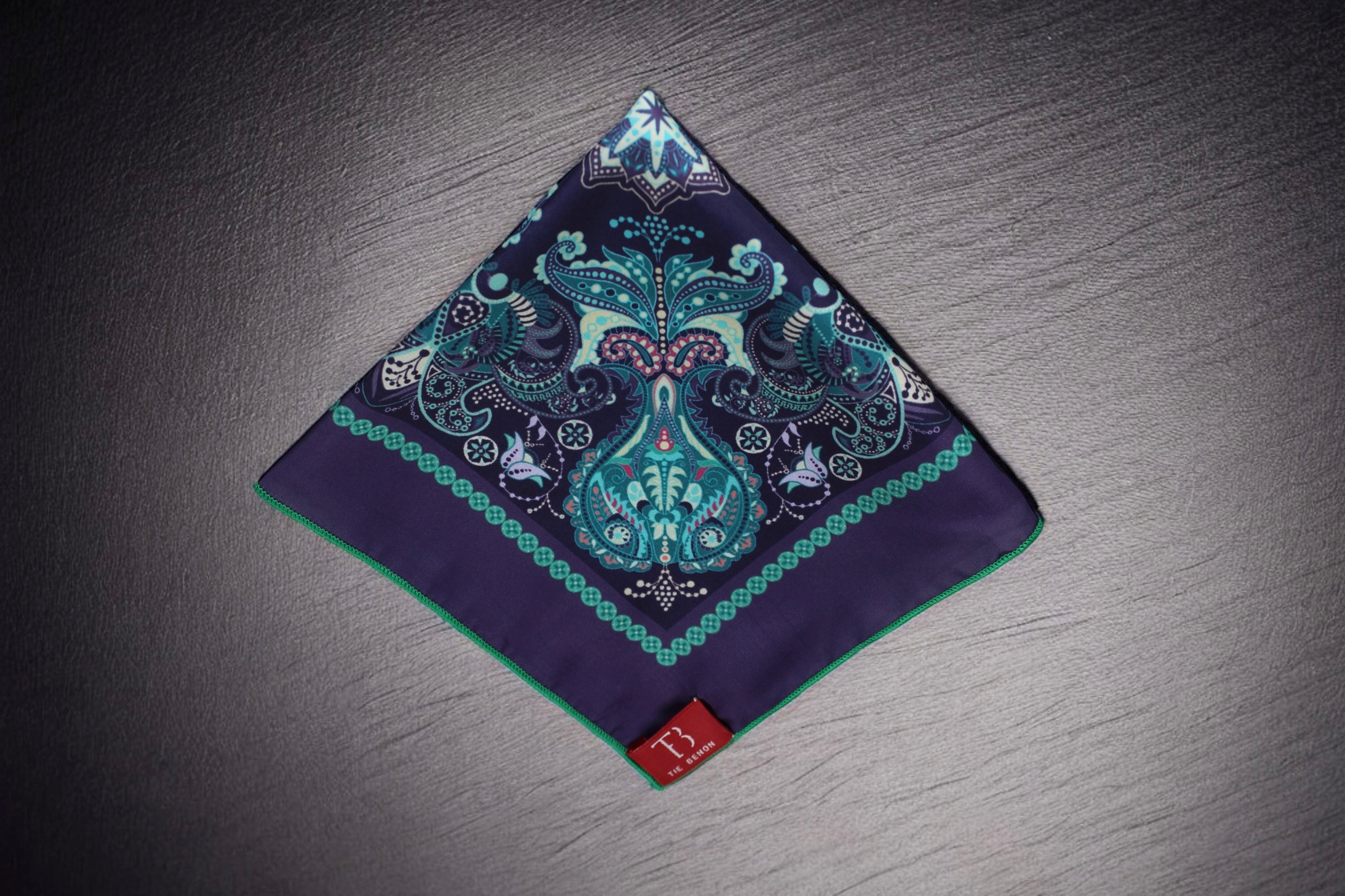 A folded navy and teal pocket square with paisley design displayed on a dark surface.