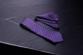 A purple geometric tie and pocket square set laid out on a dark surface.