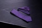 A purple geometric tie and pocket square set laid out on a dark surface.