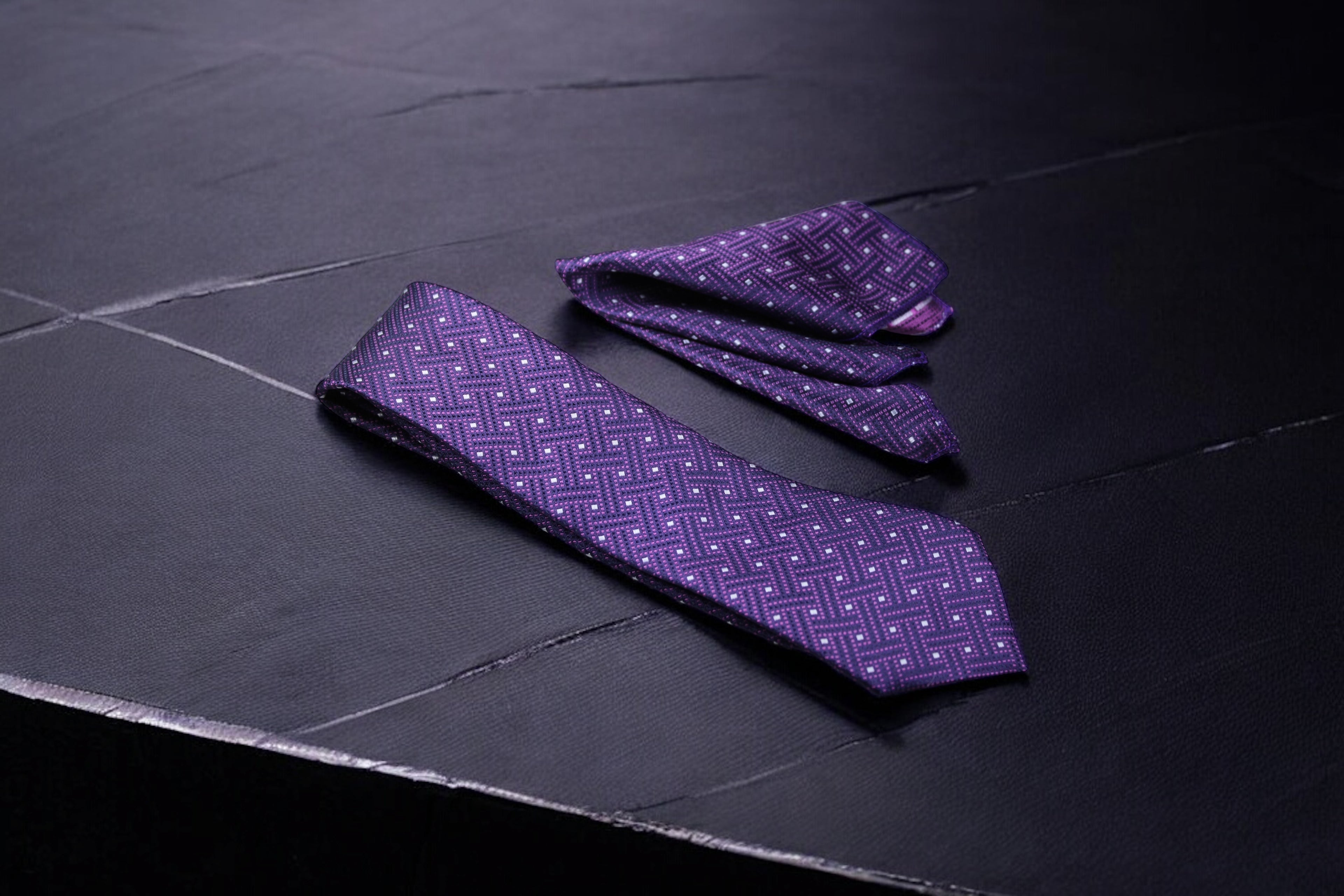 A purple geometric tie and pocket square set laid out on a dark surface.
