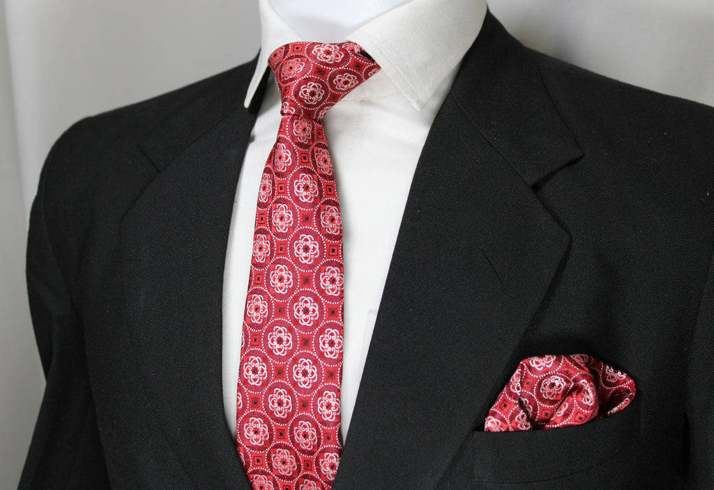 A red geometric patterned tie and pocket square set displayed on a mannequin wearing a black suit and white shirt.