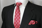 A red geometric patterned tie and pocket square set displayed on a mannequin wearing a black suit and white shirt.