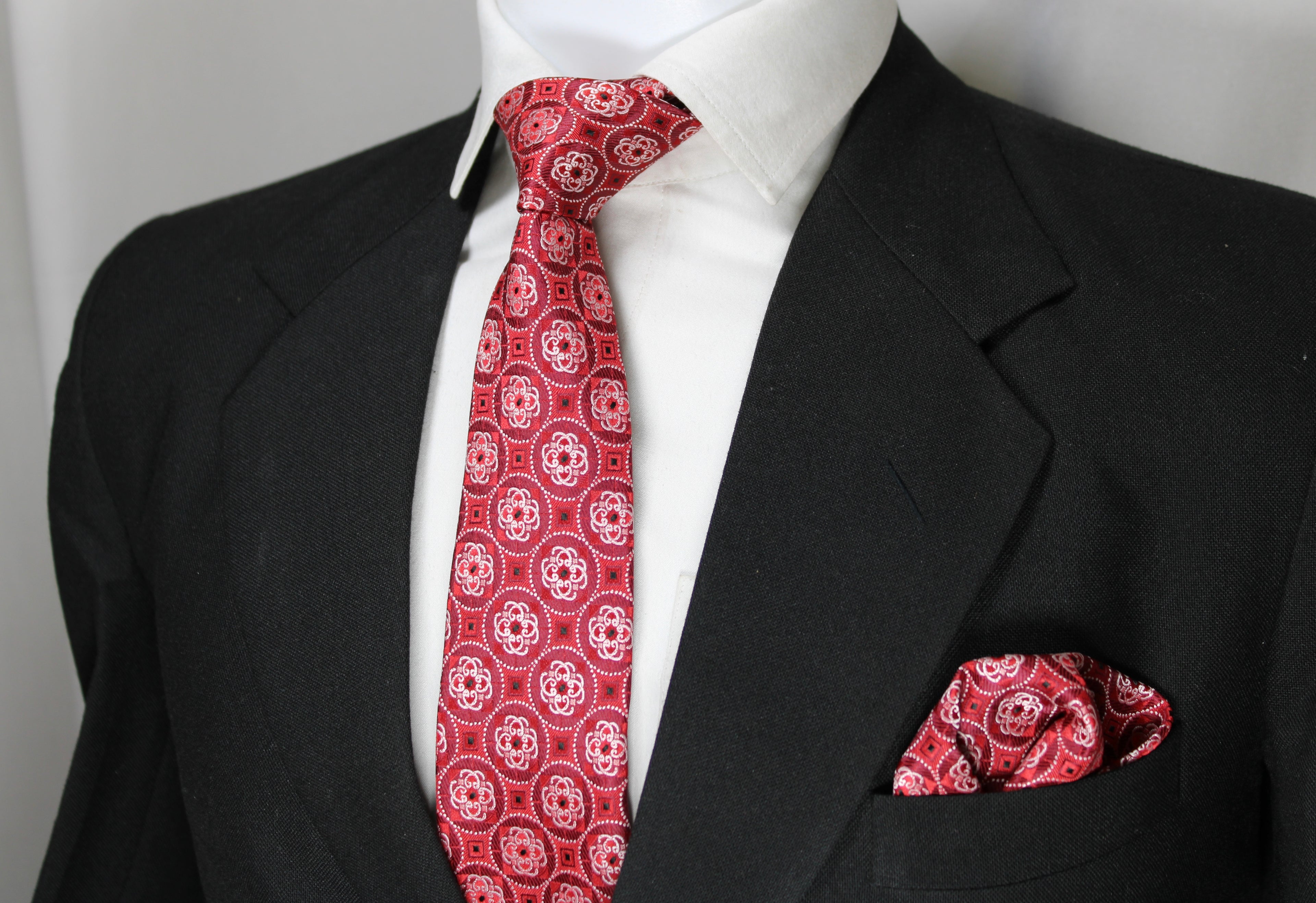A red geometric patterned tie and pocket square set displayed on a mannequin wearing a black suit and white shirt.