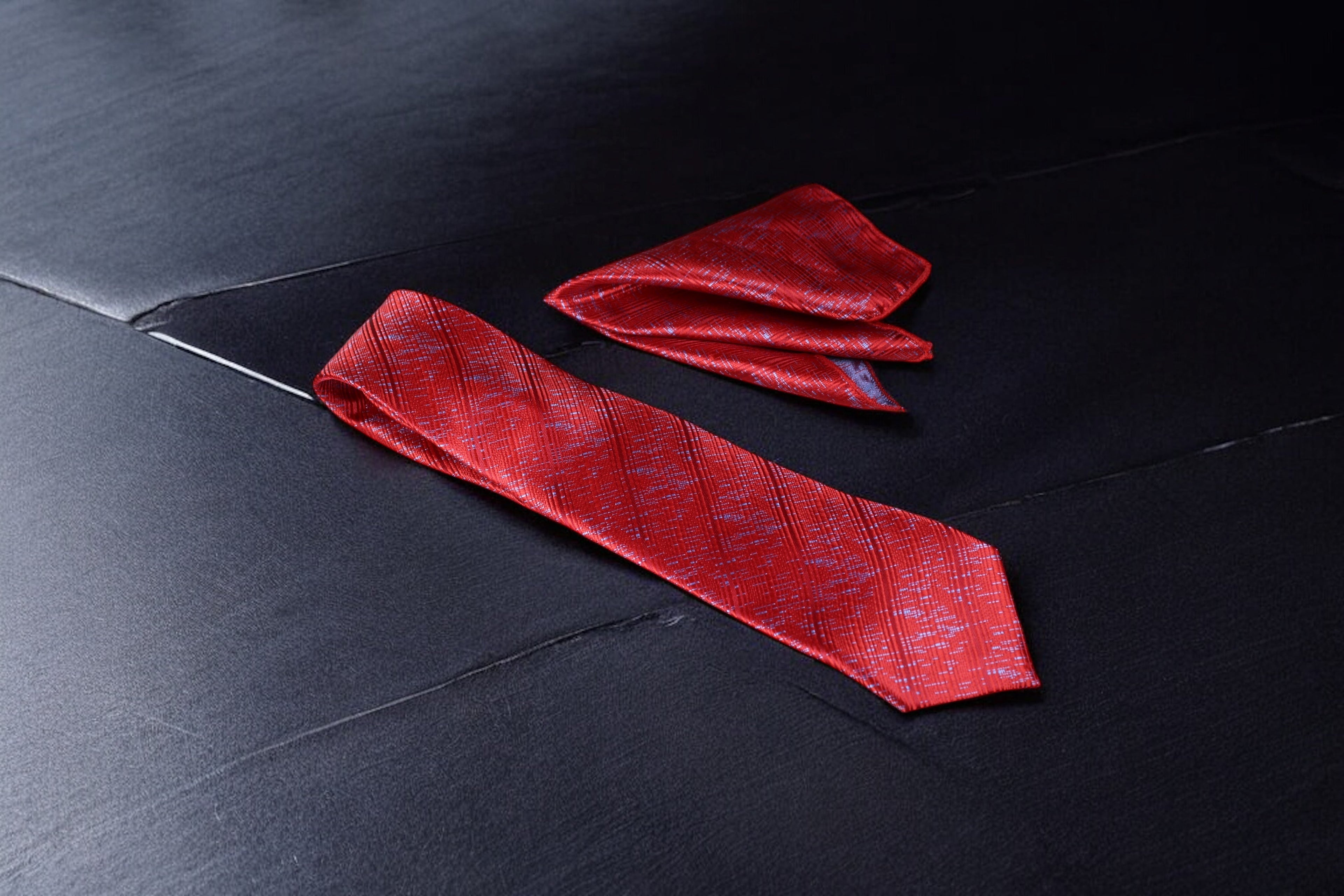 A red necktie with a pattern and a matching pocket square placed on a dark surface.