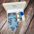 A Firozi Paisley Printed Silk Tie in a blue color with a classic paisley pattern, displayed in an open white box, alongside a matching pocket square and a brass tie pin.